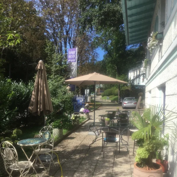 Gartenterrasse des Cafés unter alten Bäumen, Tische und Stühle im Schatten.