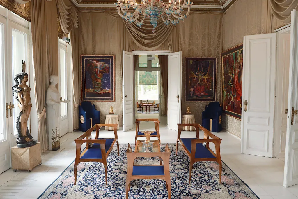 Der Große Salon der Villa: Kronleuchter, blaue Sessel um einen Glastisch, Skulpturen und Gemälde, dahinter der Blick in den Garten.