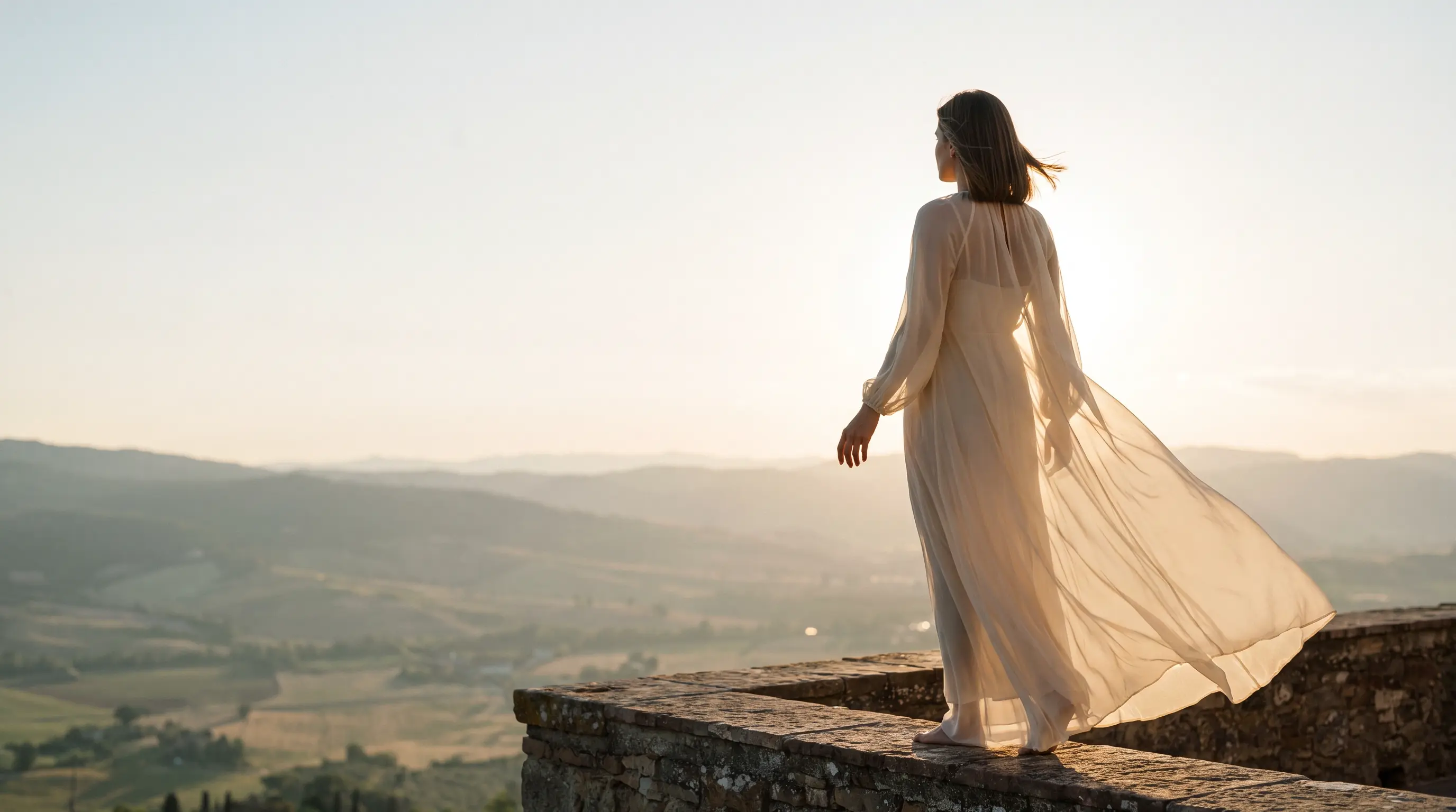 Eine Frau in fließendem Kleid steht auf einer Terrasse und blickt über hügelige Landschaft im Abendlicht.
