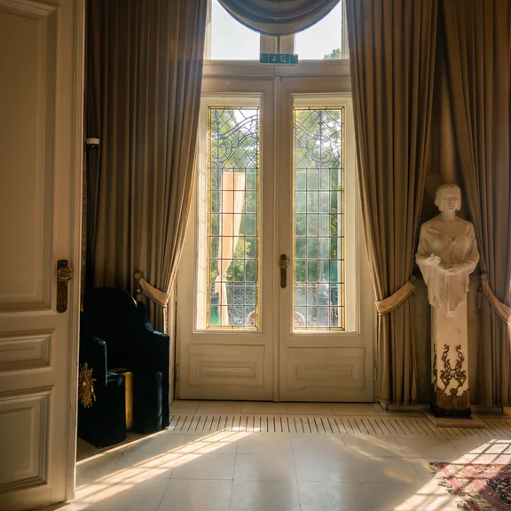 Hohe Fenstertür der Villa mit schweren Vorhängen, Nachmittagslicht auf dem Boden.