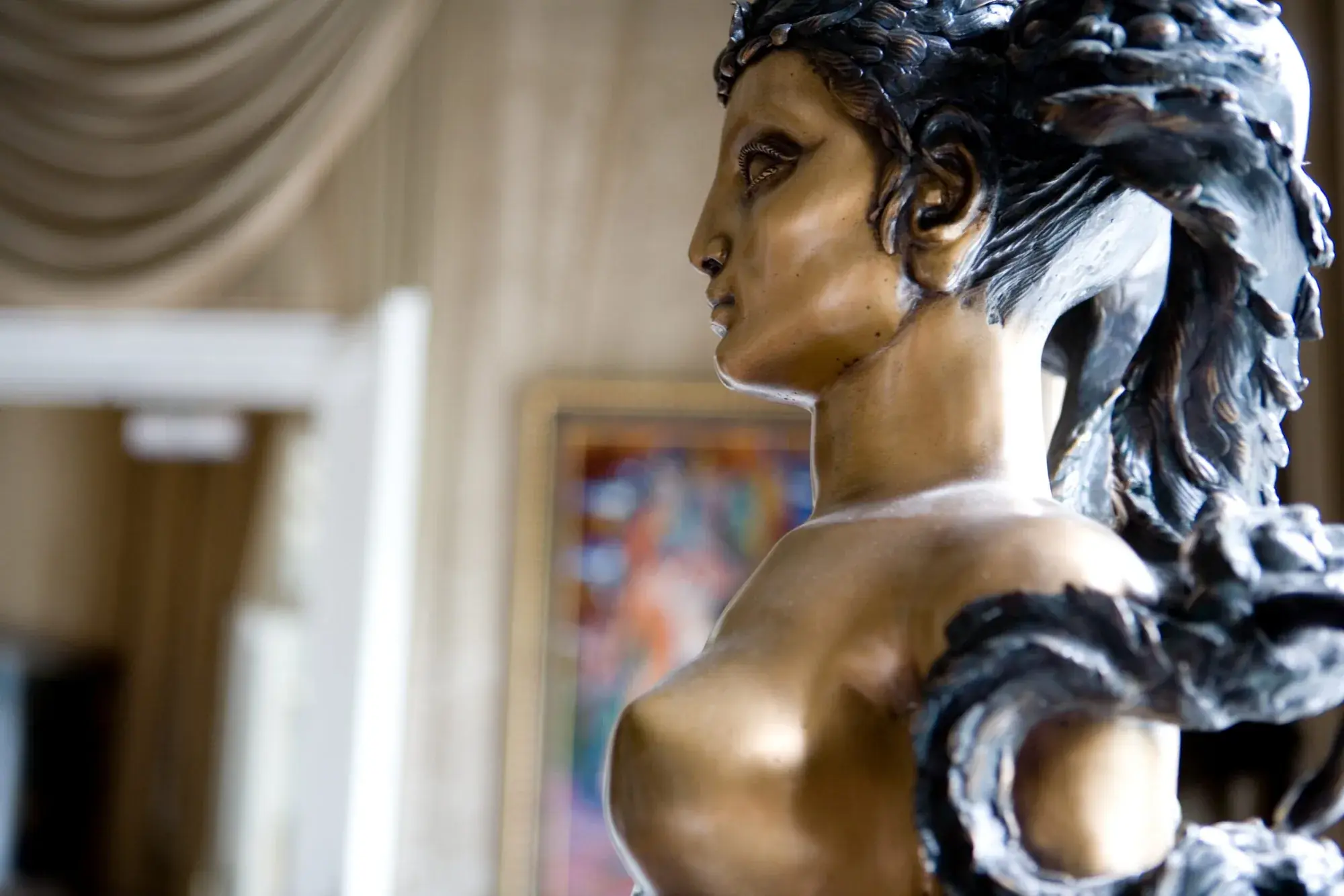 Eine Bronzefigur im Profil, dahinter unscharf ein gerahmtes Gemälde und ein heller Vorhang.